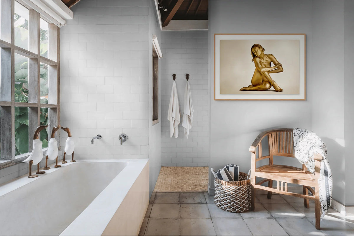 Gold fine art photograph by Zack Zublena of a seated woman painted in gold, framed in light oak above a white bathroom wall, with a teak chair, a woven basket and natural light in a luxury spa bathroom