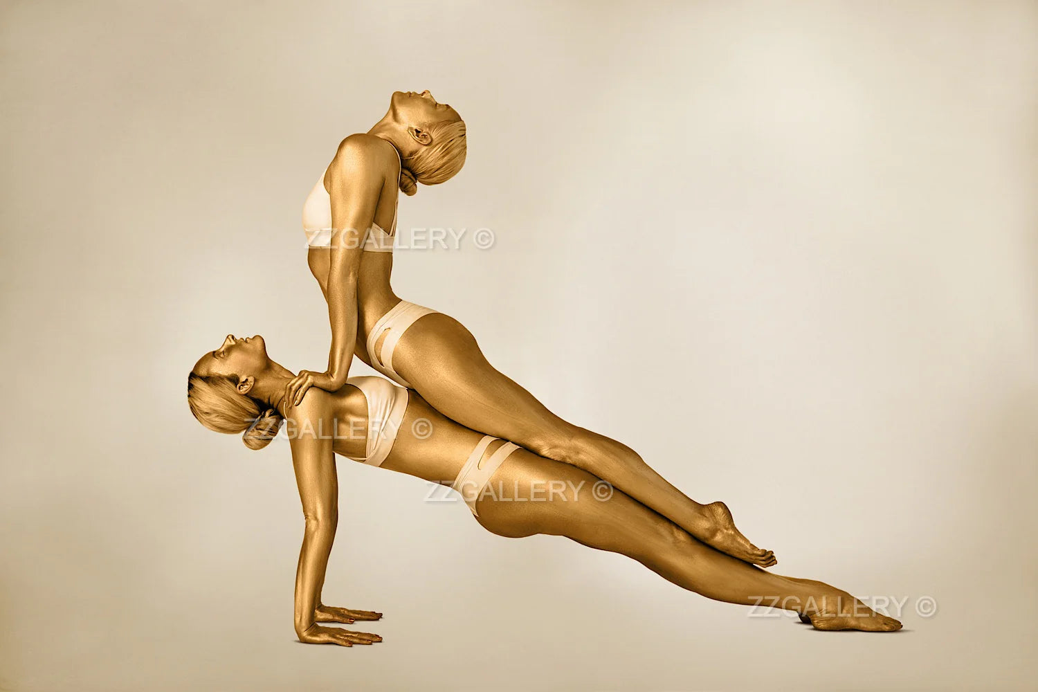 Golden-painted female duo in balance acrobatic pose, fine art photography on museum paper, luxury decor for hotels, spas, and collectors.