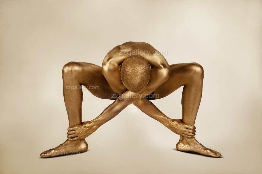 Golden-painted figure in a deep squat pose, arms crossing and gripping ankles, representing strength, meditation, and edgy conceptual fine art photography.