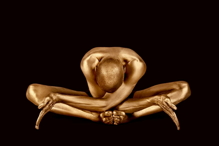 Golden-painted male figure in a deep yoga stretch with head bowed forward, captured against a black background. Fine art photography by Zack Zublena for luxury wall décor in modern interiors.