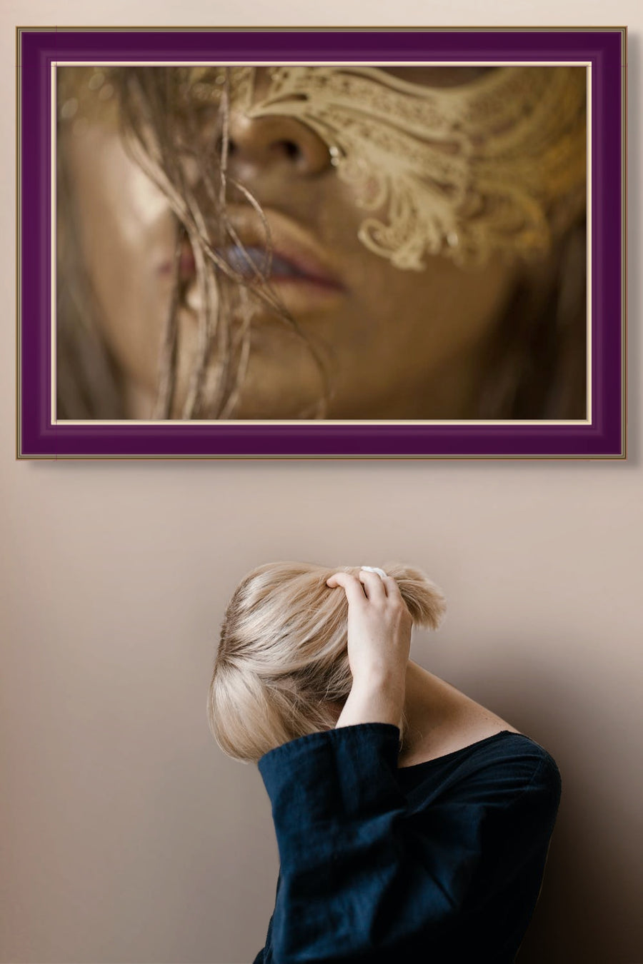Golden masked portrait artwork in a luxurious purple frame displayed on a modern wall above a woman with blonde hair, elegant fine art wall decor.