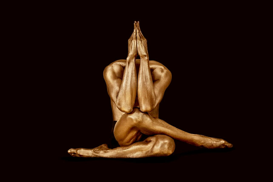 Golden male figure seated with legs crossed and arms extended upward in a prayer-like yoga pose, fine art photography against a deep dark backdrop.