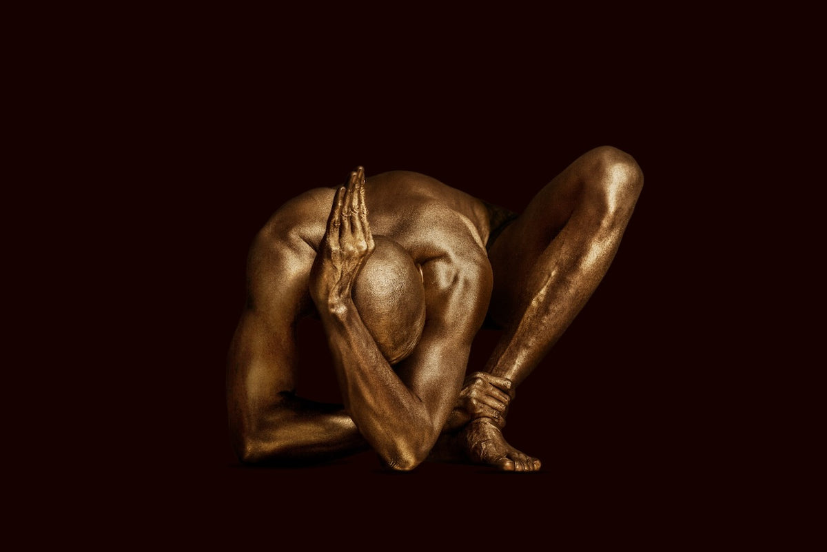 "Salutation to the Sun" by Zack Zublena: a fine art photograph of a gold-painted male figure bowed low in the dark, hands joined in prayer like a flame, before the sun has risen. Limited edition gold body paint photography, dark background.