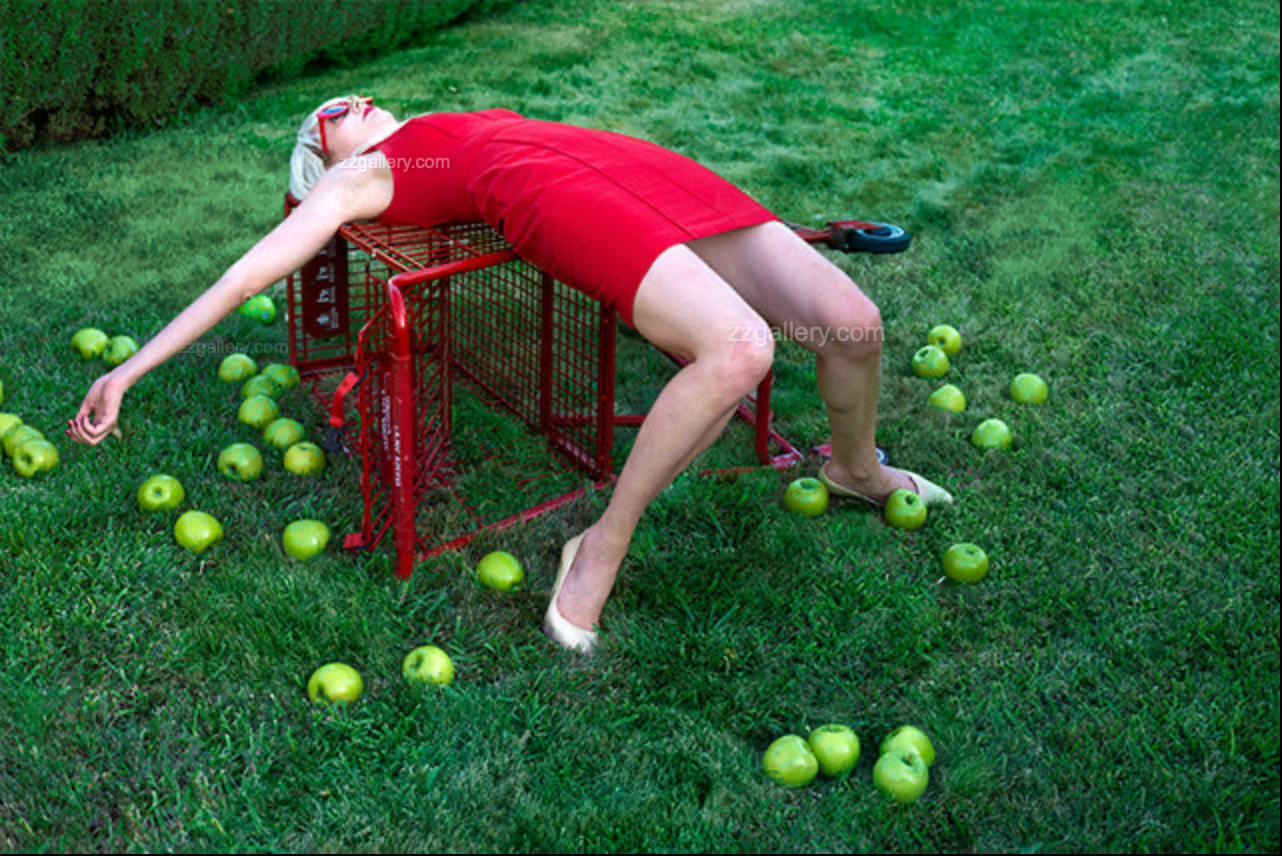 Surreal pop art photograph of a woman in a red dress sprawled across an overturned shopping cart in a grassy garden, surrounded by scattered green apples.