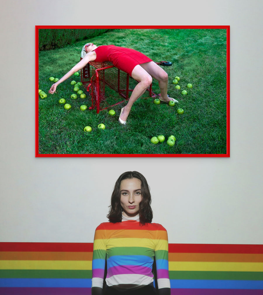 Surreal pop art photograph of a woman in a red dress stargazing while sprawled over a red shopping cart on grass, surrounded by apples – whimsical fine art print.