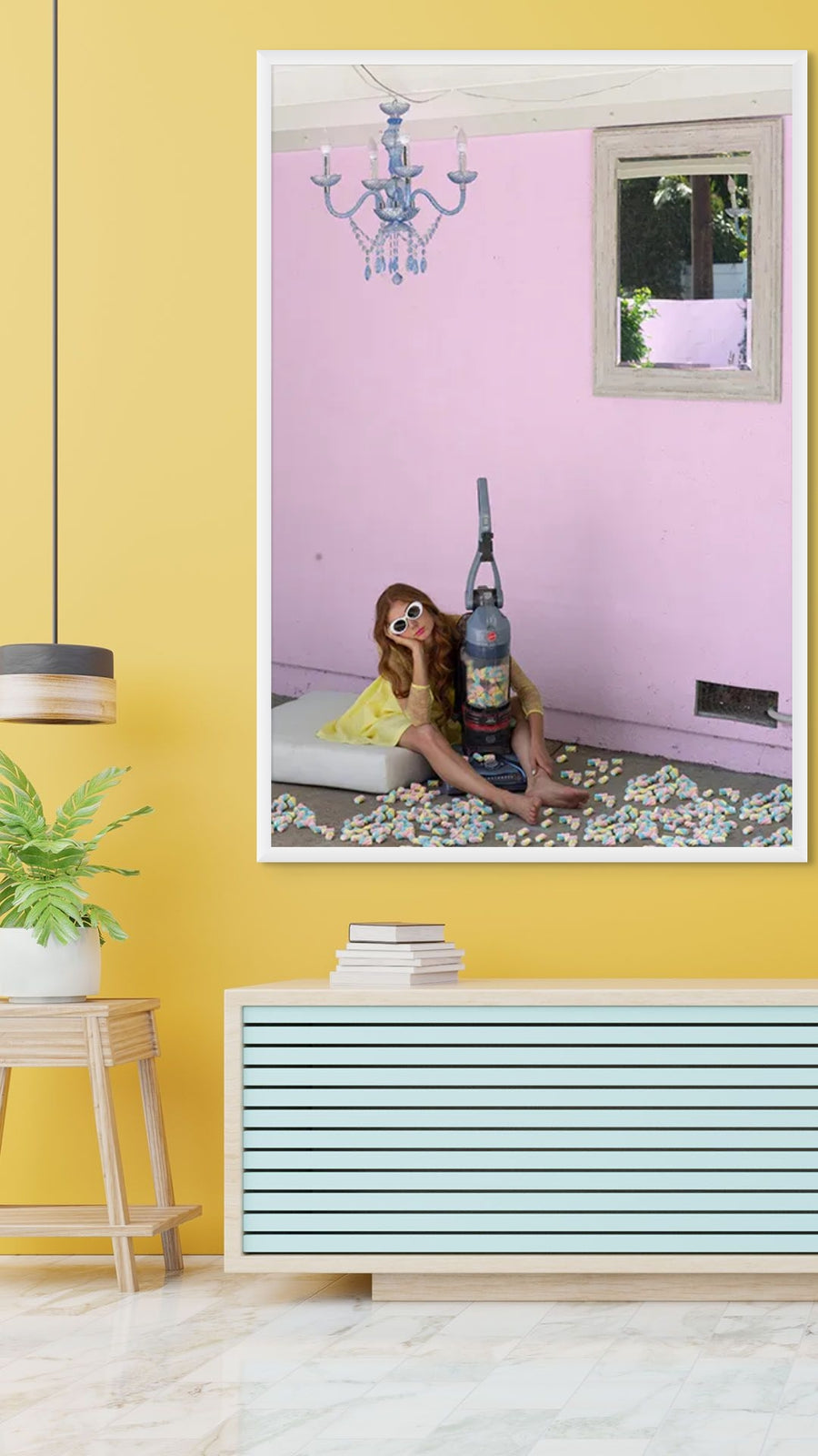 Whimsical fine art photograph of a woman in a yellow dress sitting beside a vacuum cleaner filled with candy, set against a pink wall – surreal wall art for playful interiors and modern home décor.