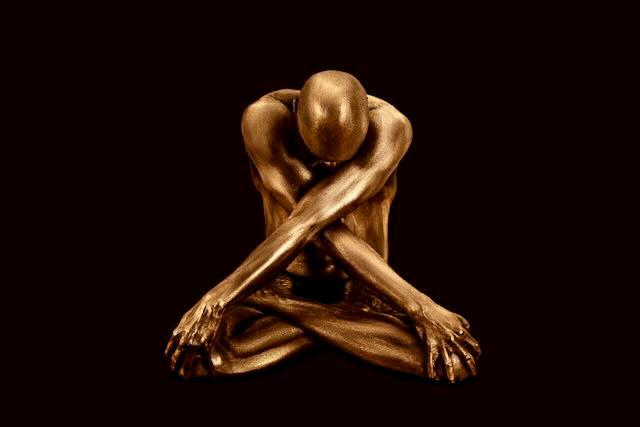 Gold-painted figure seated, head bowed, arms crossed in an X over the knees, against a deep black background. Fine art photography by Zack Zublena.