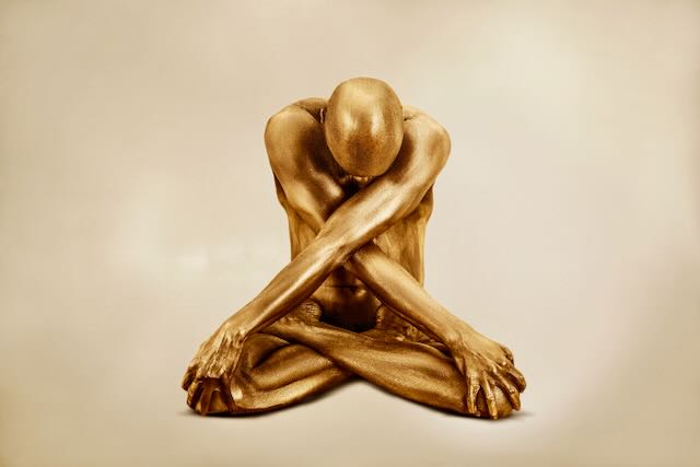 I Refuse to Perform by Zack Zublena: gold-painted figure seated, head bowed, arms crossed in an X over the knees, on warm cream.