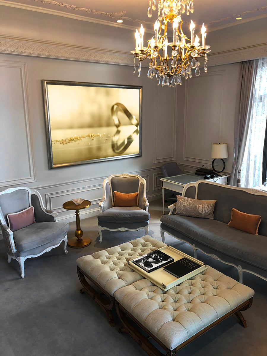 Elegant fine art photograph of a golden ring casting a heart-shaped shadow with scattered gold dust, displayed in a luxurious living room interior with chandelier and classic furniture.