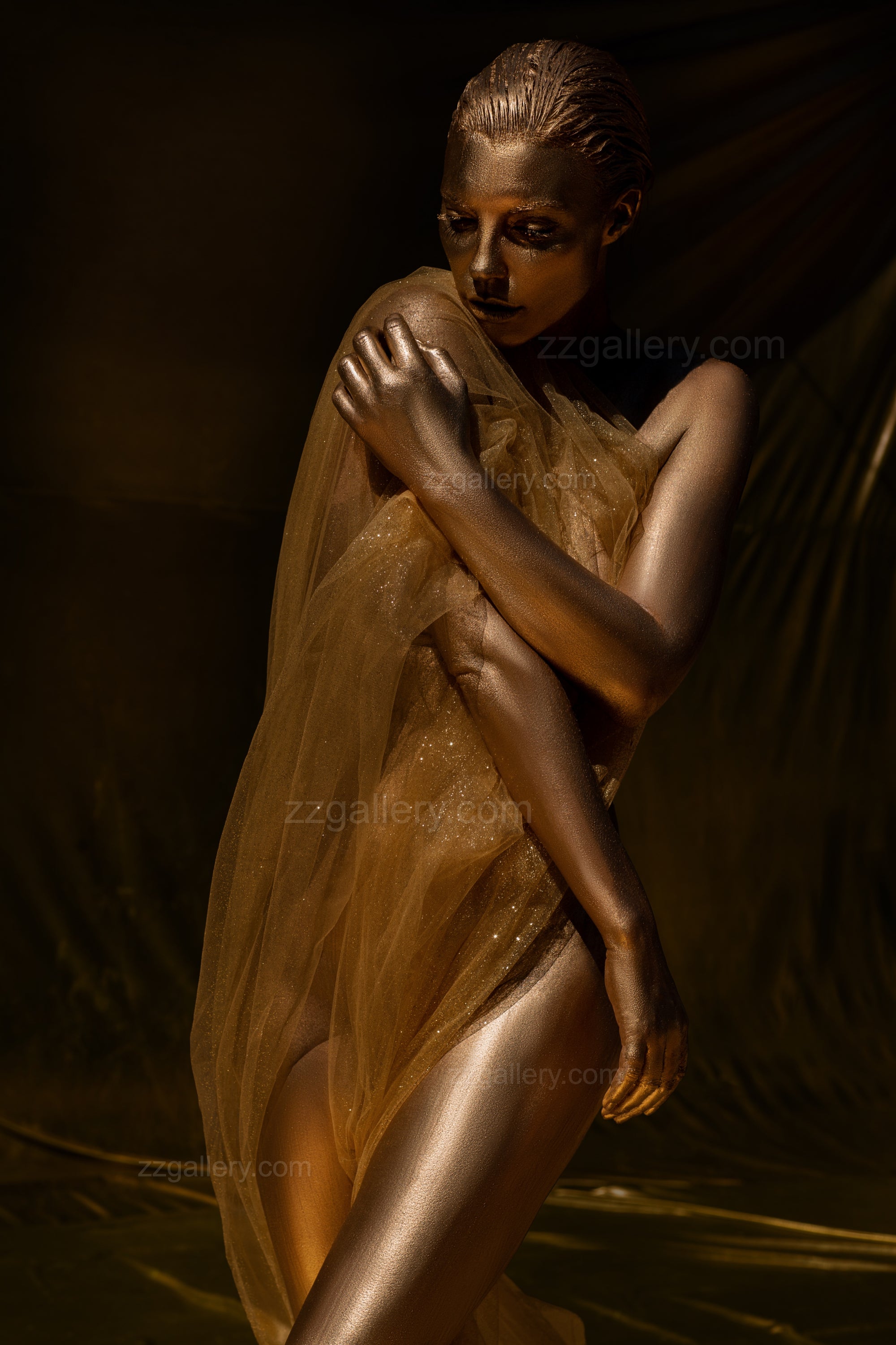 Golden fine art portrait of a woman with metallic body paint, draped in sheer fabric, posing with arms crossed over her chest.