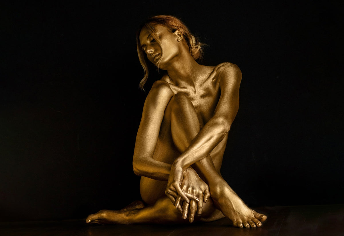 Moonlit Reverie III – Surrender by Zack Zublena: gold-painted woman seated with her arms around her knee and hands clasped against a dark background, from the Dark Elegance collection