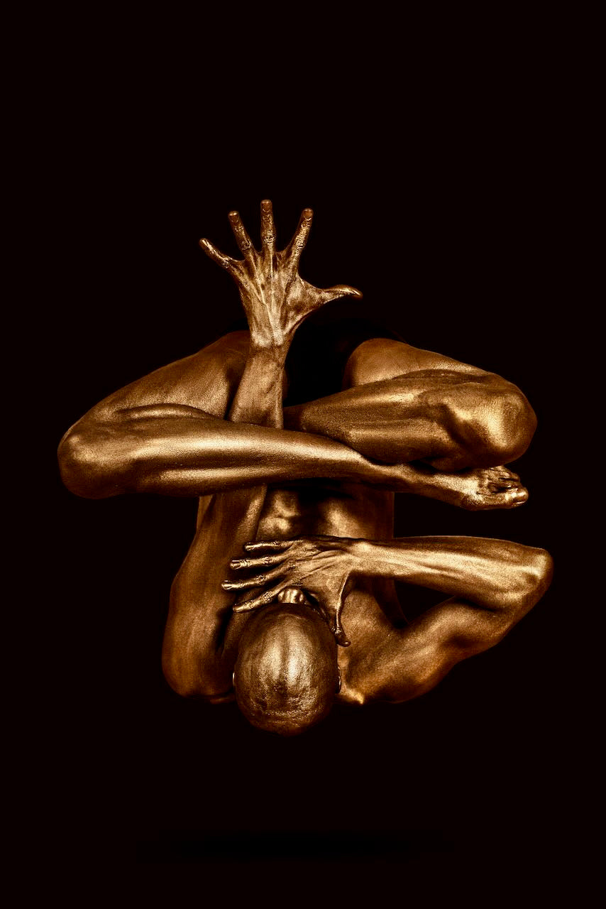 Golden-painted figure in a sculptural yoga pose, one arm extended upward with open hand, set against a deep dark background — moody fine art photography suitable for luxury interiors and modern spaces."