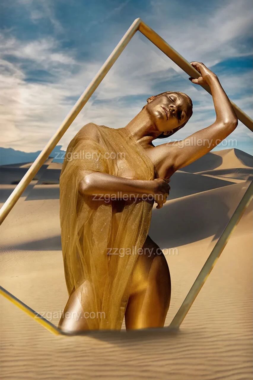 Golden-painted nude woman draped in sheer fabric posing in a desert landscape with modern geometric frame