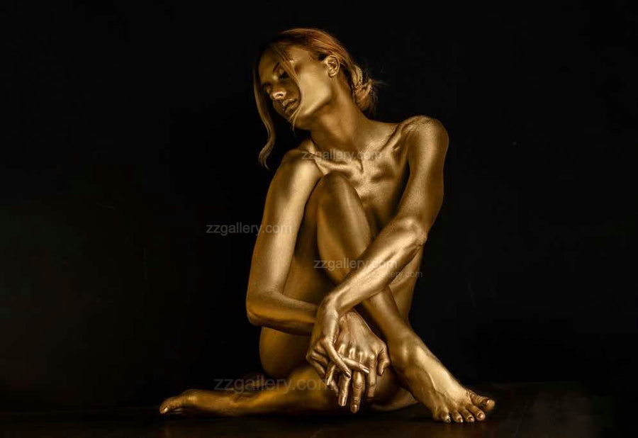Framed fine art photograph of a golden-painted figure seated gracefully with arms wrapped around her knees, displayed in a luxury modern interior with dark wood cabinet and minimalist décor. Contemporary wall art for upscale living rooms, hotels, or galleries.