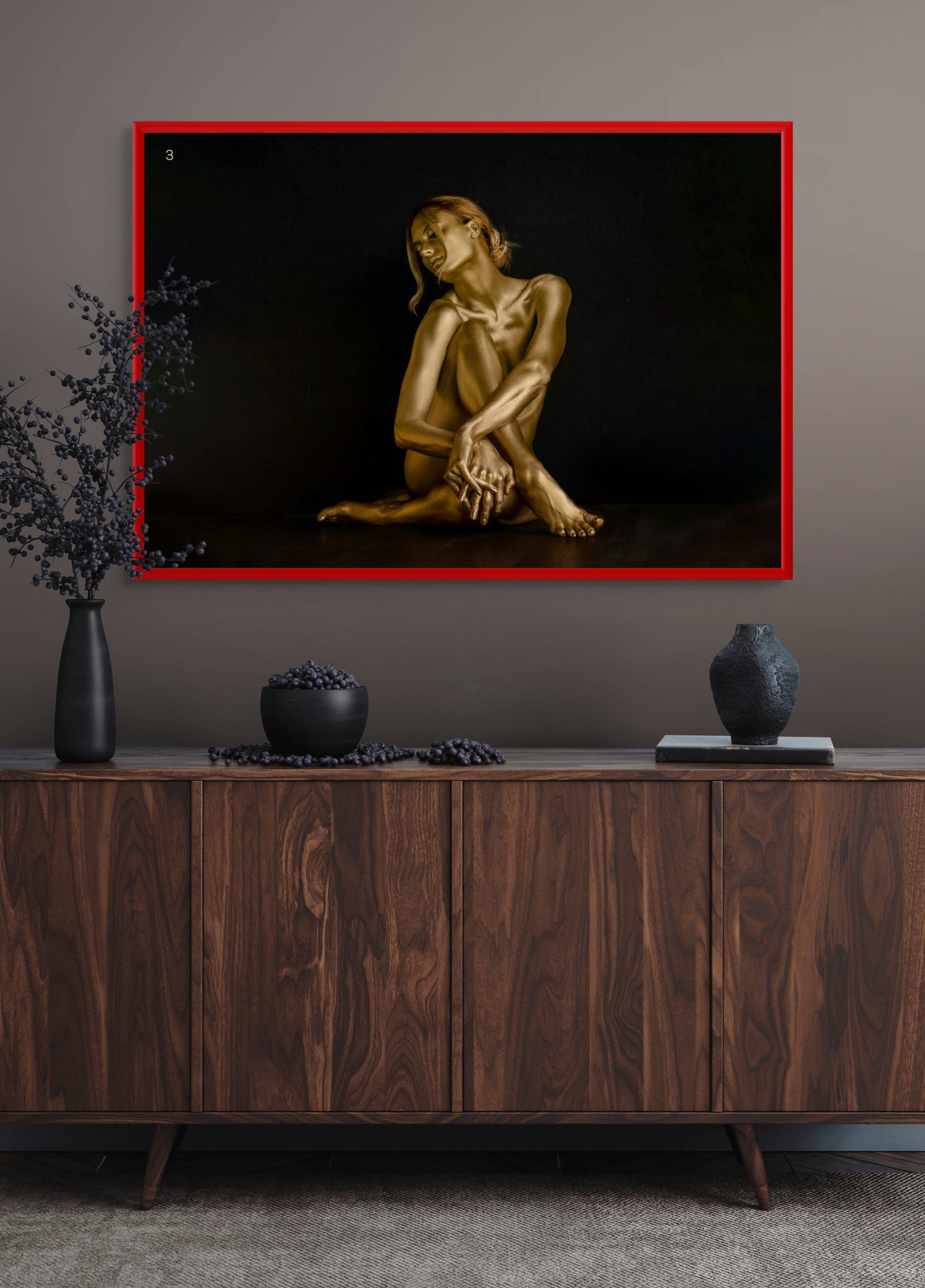 Framed fine art photograph of a golden-painted figure seated gracefully with arms wrapped around her knees, displayed in a luxury modern interior with dark wood cabinet and minimalist décor. Contemporary wall art for upscale living rooms, hotels, or galleries.