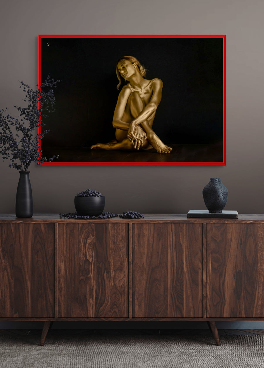 Framed fine art photograph of a golden-painted figure seated gracefully with arms wrapped around her knees, displayed in a luxury modern interior with dark wood cabinet and minimalist décor. Contemporary wall art for upscale living rooms, hotels, or galleries.