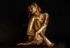 Golden-painted nude woman seated gracefully with legs folded and arms crossed, set against a dark black background. Fine art photography highlighting feminine elegance, sensuality, and sacred beauty, ideal for luxury interiors, spas, and contemporary art collections.