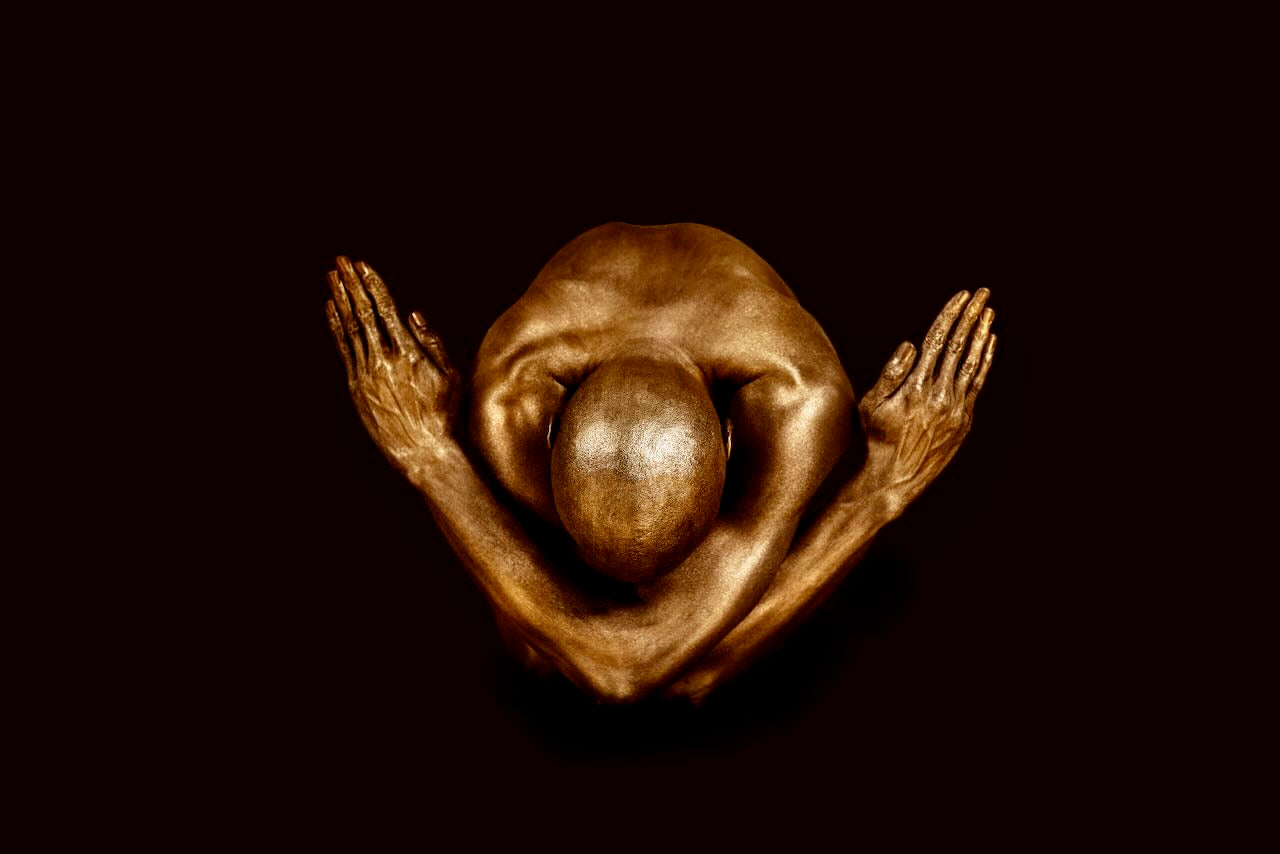 Golden-painted male figure in a powerful crossed-arms yogi pose, seated against a deep black background. Fine art photography for moody interiors and meditation spaces.