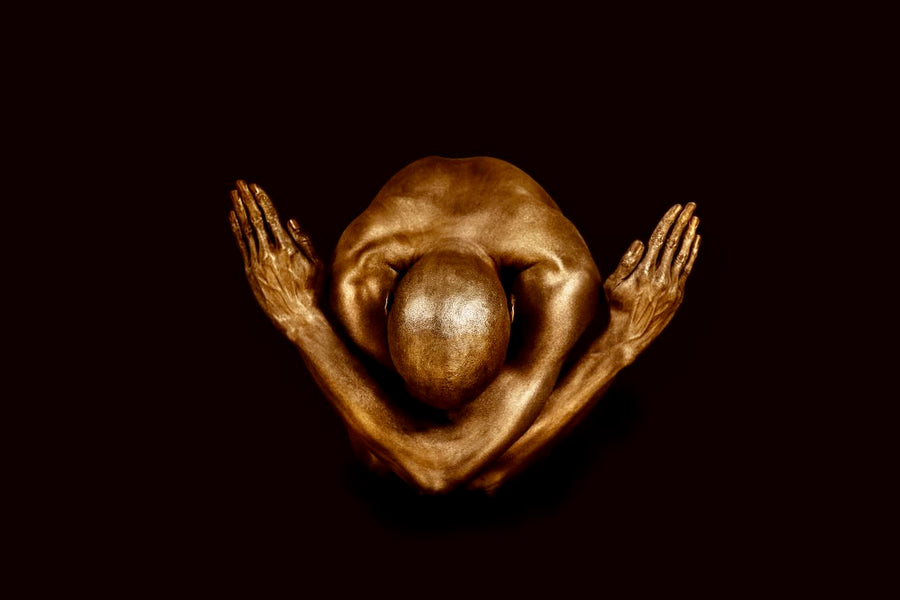 Golden-painted male figure in a crossed-arms yogi pose against a deep dark backdrop, framed as fine art wall decor in a modern luxury interior.