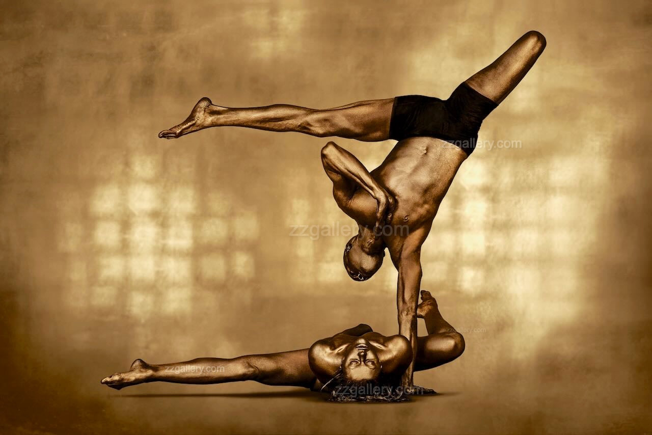 Golden-painted couple performing an acrobatic balance pose, man lifted in a one-arm handstand over woman’s body, contemporary fine art photograph by Zack Zublena.