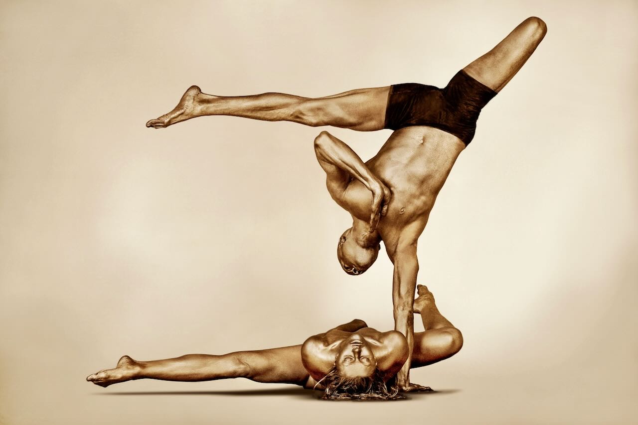 Golden-painted couple performing an acrobatic yoga balance pose, fine art photography by Zack Zublena. Luxury wall art suitable for yoga studios, spas, hotels, and modern interiors.
