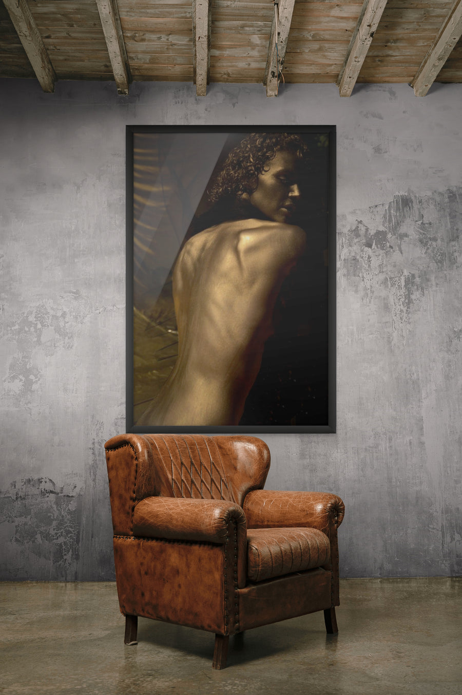 Golden-painted figure photographed from the back, displayed in an industrial-style mockup with a leather vintage armchair and concrete wall.