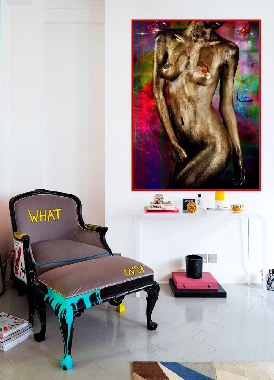 Golden-painted figure artwork with butterflies on a vibrant multicolor background, displayed as framed wall art in a modern eclectic interior with a bold accent chair.