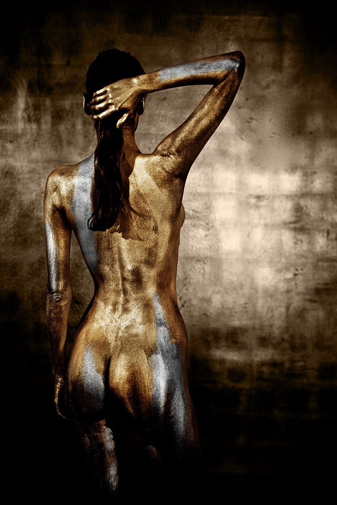 Golden-painted nude female back fine art photograph displayed in a rustic bohemian living room with wicker armchair and earthy tones.