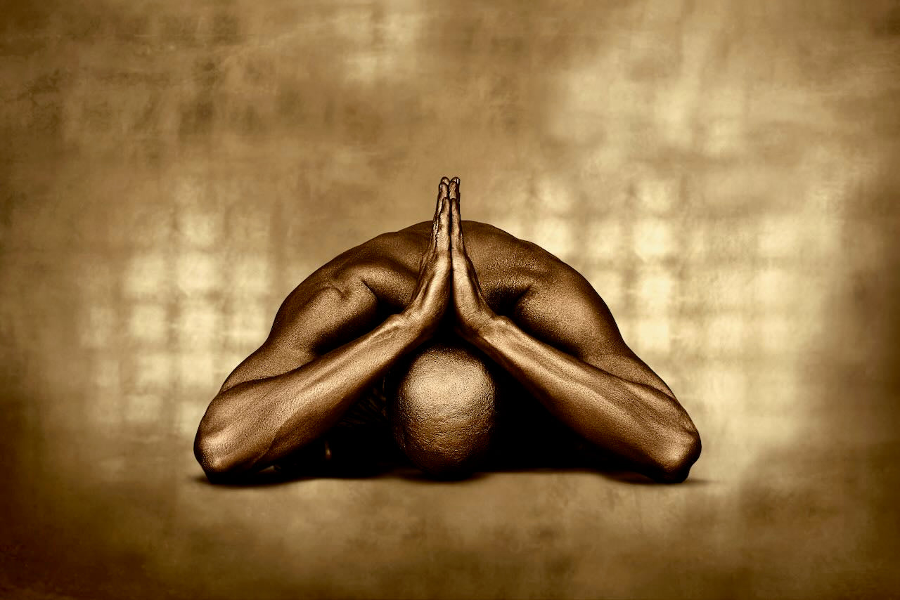 Gold-painted male figure with folded hands in a serene pose, fine art photography on a radiant golden background.