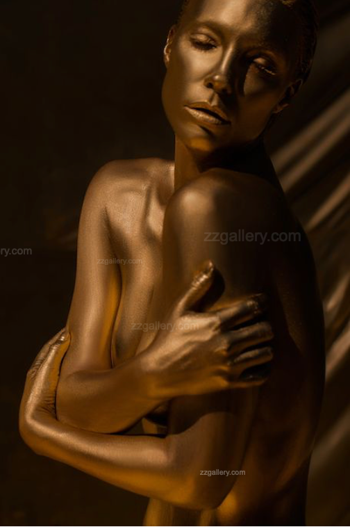 Elegant golden-painted nude woman with eyes closed and arms wrapped around her shoulders, captured in warm light against a neutral background. Contemporary fine art nude photography symbolizing serenity, self-love, and feminine strength — ideal for luxury interiors, spas, and collector wall art.