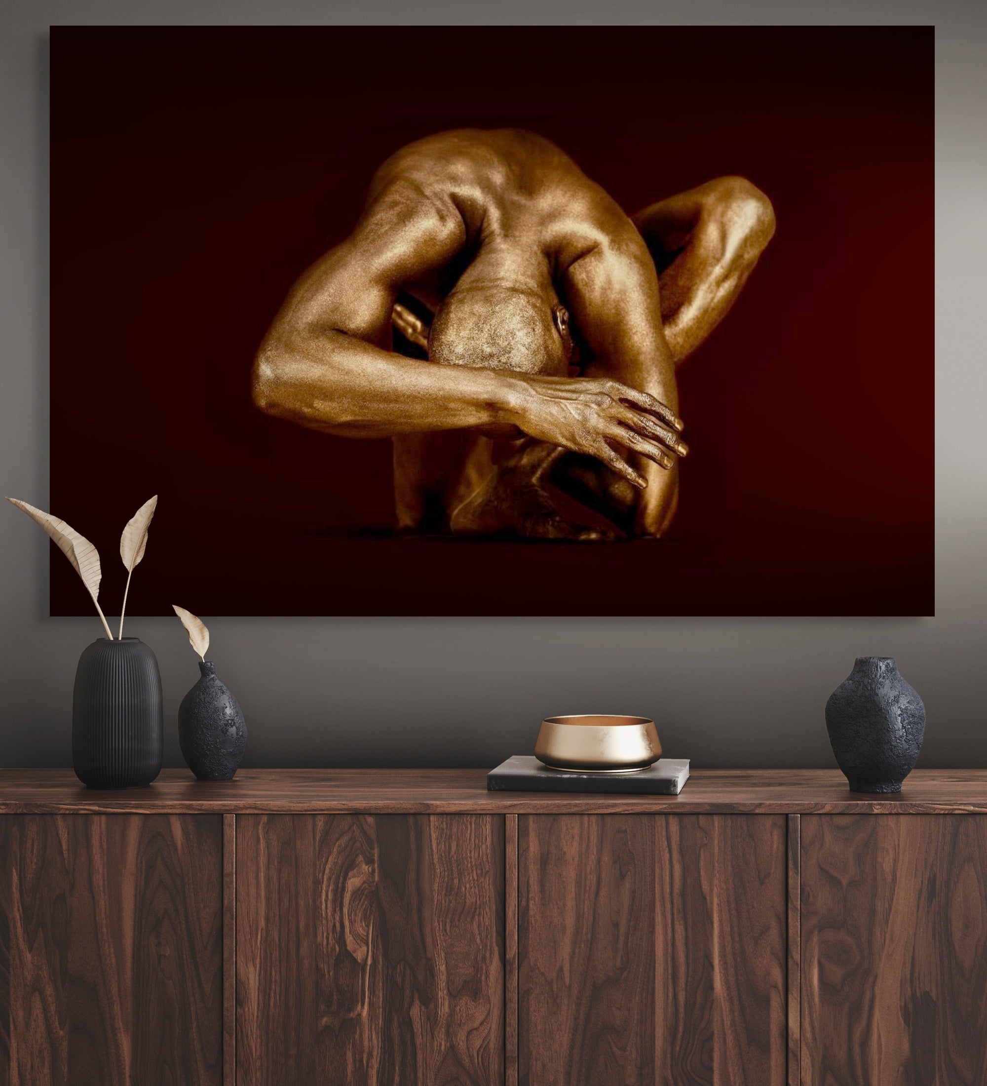 Golden-painted male figure in a sculptural contemplative yoga pose, fine art wall decor on a deep red background above a modern wooden console.