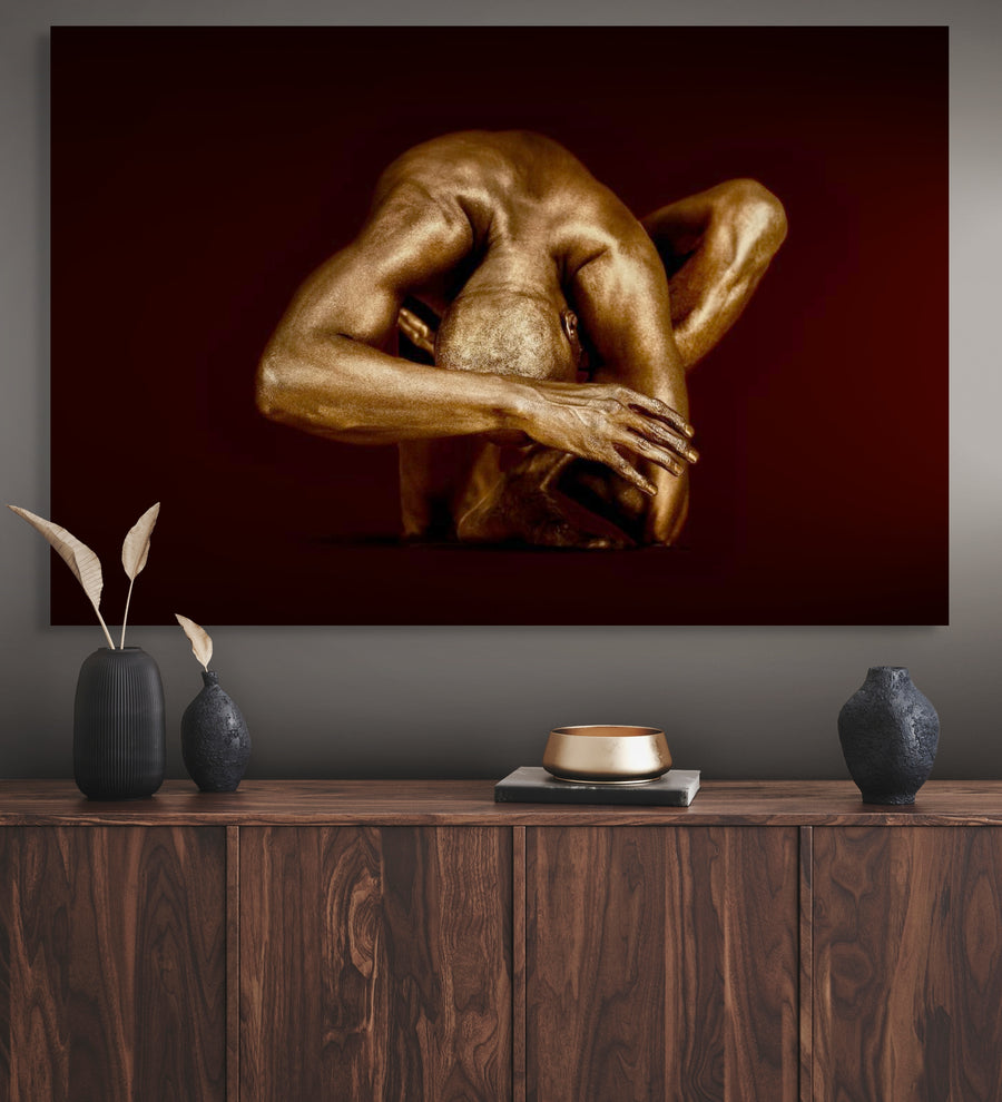 Golden-painted male figure in a sculptural contemplative yoga pose, fine art wall decor on a deep red background above a modern wooden console.