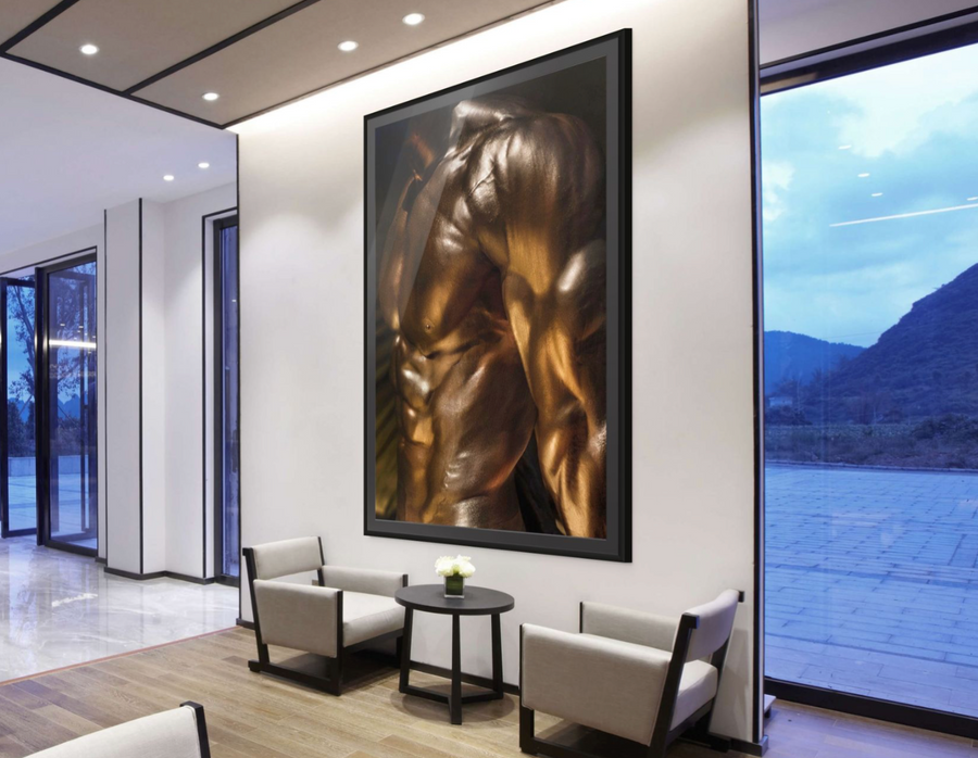 Framed golden fine art photograph of a muscular male torso with metallic body paint, displayed in a modern lobby with armchairs, table, and large windows overlooking mountains.