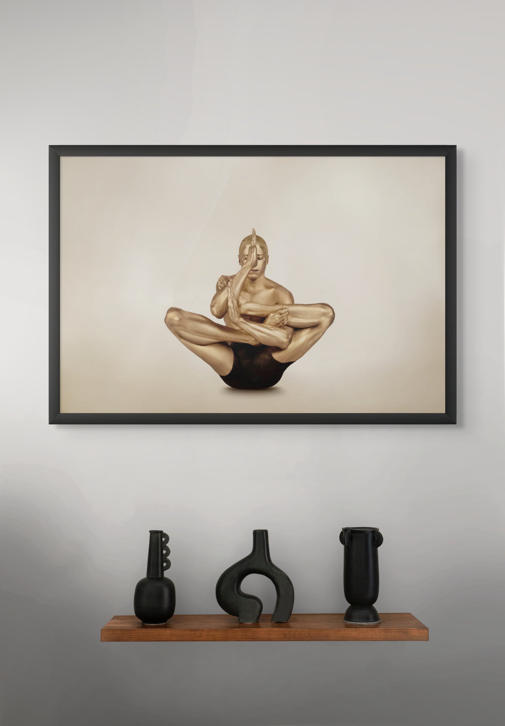 Golden-painted male figure in advanced yoga pose, fine art photograph displayed as framed wall art above a modern wooden shelf with black vases.