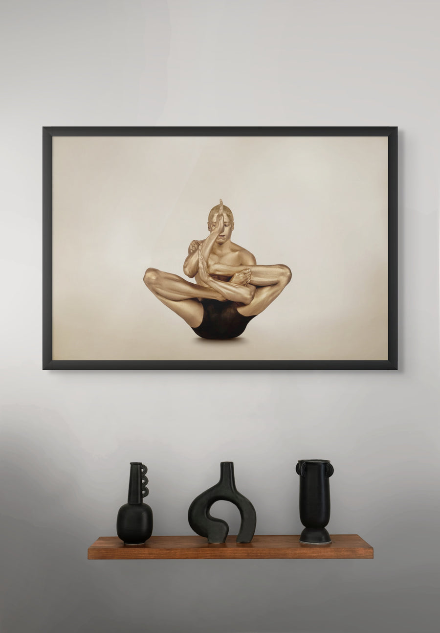 Golden-painted male figure in advanced yoga pose, fine art photograph displayed as framed wall art above a modern wooden shelf with black vases.