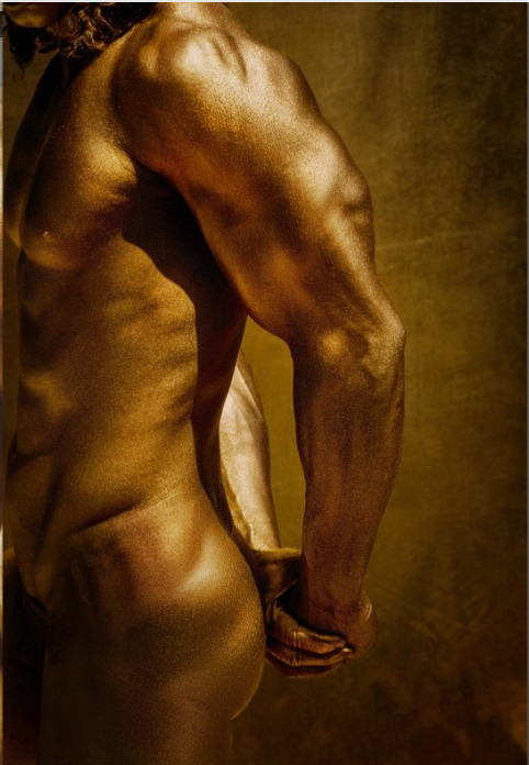 Golden male nude artwork by Zack Zublena – a close-up of a sculpted masculine back and arms painted in shimmering gold, displayed in a modern interior with a wooden desk and elegant lamp.