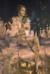 Golden-painted nude woman seen through blurred leaves and reflections, evoking mystery, softness, and ethereal femininity.