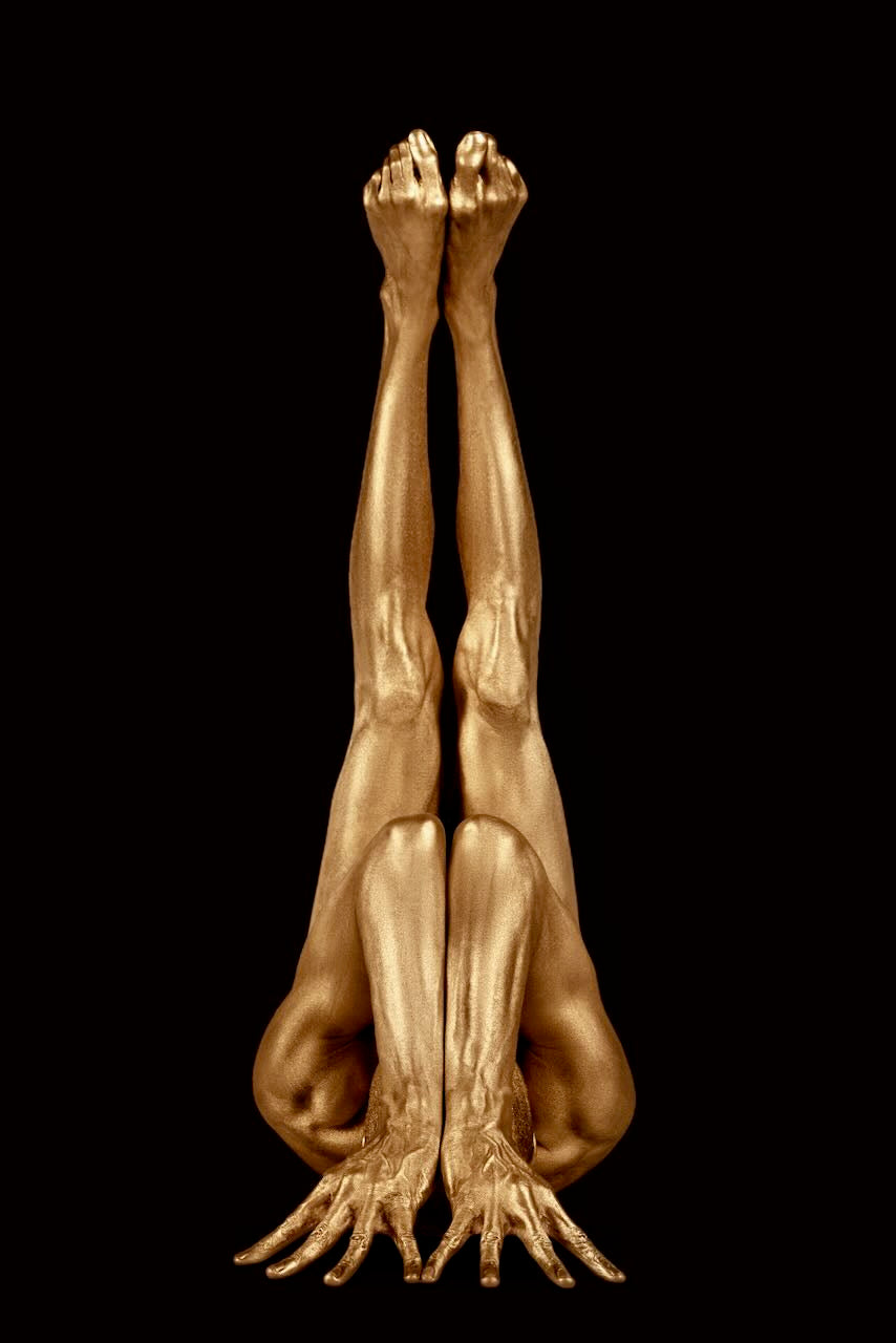 Gold-painted yogi in upside-down yoga pose with hands rooted like branches, on dark velvet background, emphasizing strength and balance.