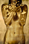 Golden nude woman blowing luminous golden particles from her hands in a shadowed setting, evoking sacred transmission and mystery.
