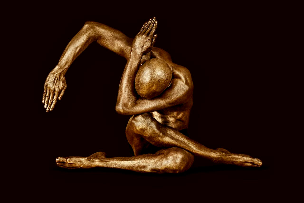 Golden-painted male figure in an abstract seated yoga pose with extended arm and bowed head, fine art photography by Zack Zublena from the Dark Elegance collection.
