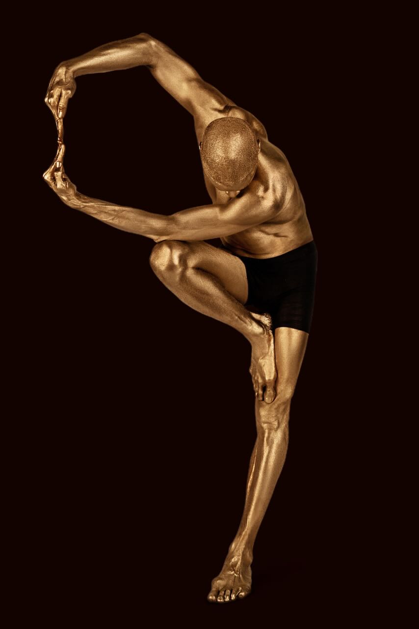 Golden-painted male dancer in a sculptural yoga balance pose, fine art wall decor displayed in a modern arched interior with leather chair and woven rug.