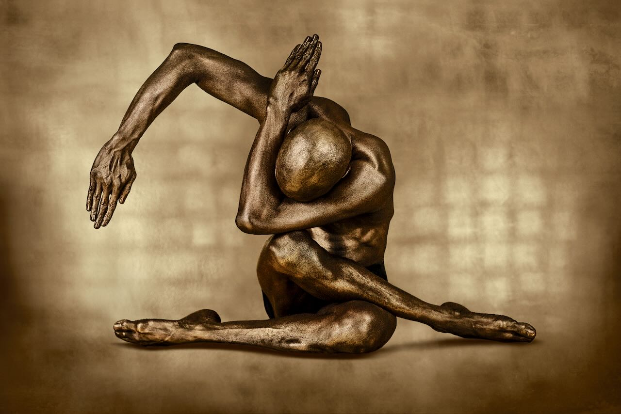 Golden-painted male figure in a seated stretch yoga pose with arm extended and head bowed, fine art photography by Zack Zublena on a luminous light background.