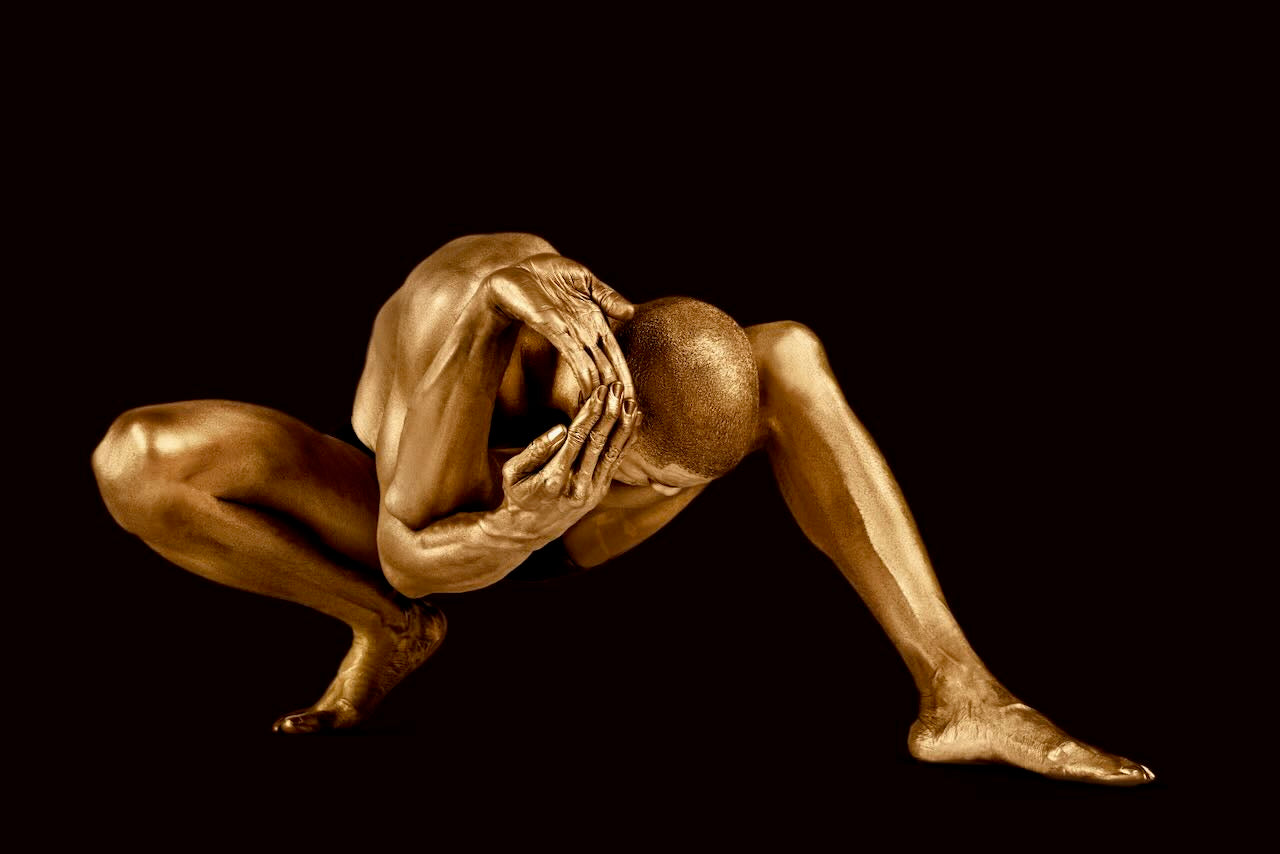 Golden-painted male figure in a crouched yoga power pose, fine art body photography in radiant gold tones on dark background.