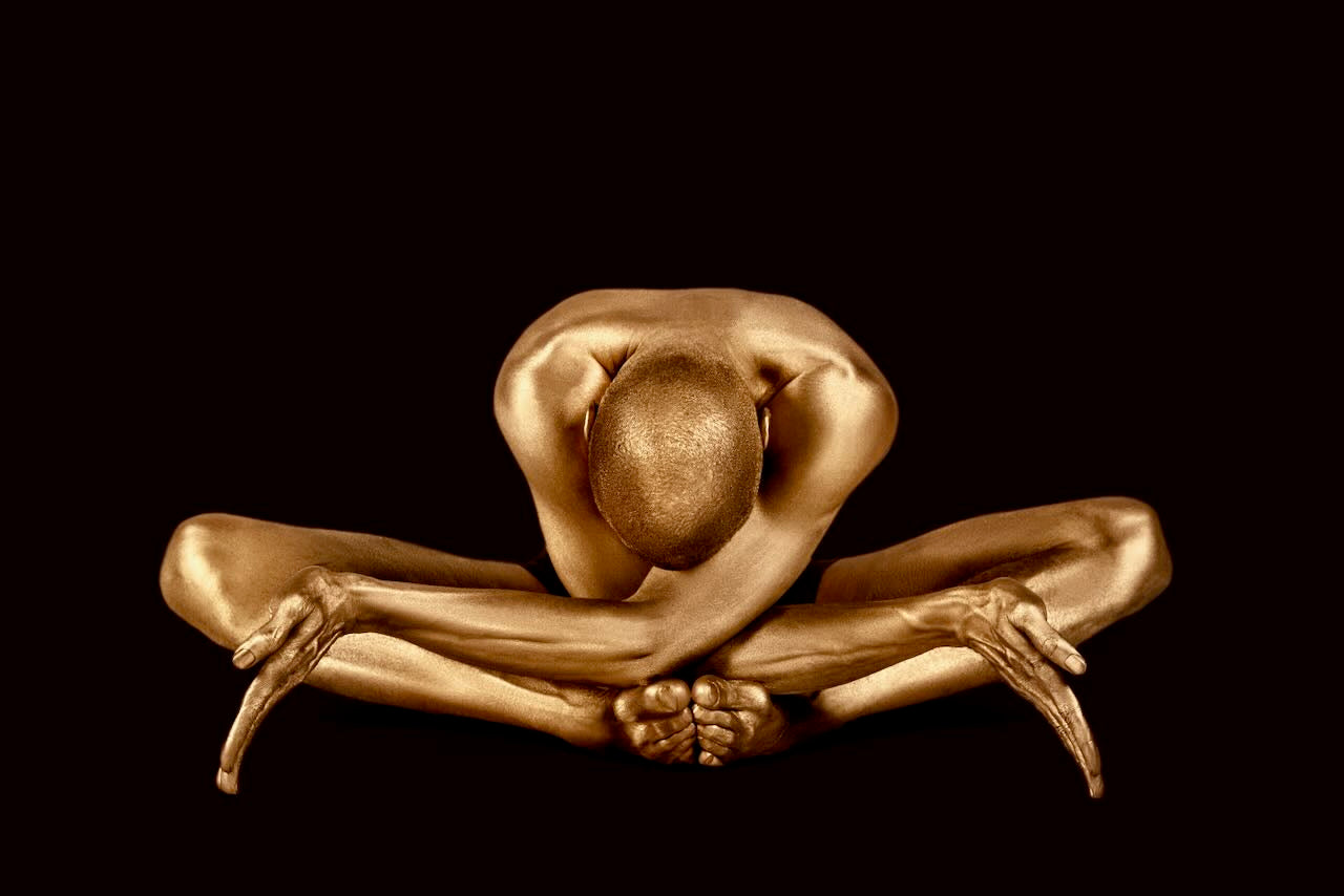 Golden-painted yogi in a forward bend pose on a deep dark background, expressing serenity, balance, and artistic elegance. Fine art wall décor by Zack Zublena.