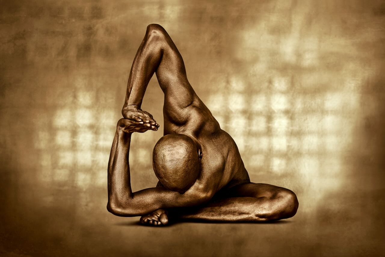 Golden-painted figure in a sculptural side bend with arm extended overhead, captured against a gold-toned background. Fine art photography highlighting strength, balance, and contemporary elegance.