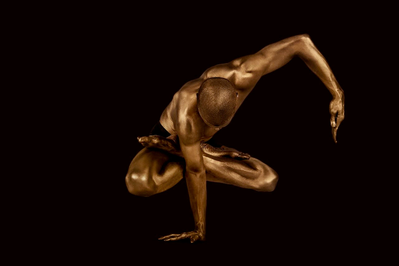 Fine art photograph of a golden-painted male figure holding a dramatic yoga balance pose against a dark background, symbolizing strength, discipline, and inner harmony.