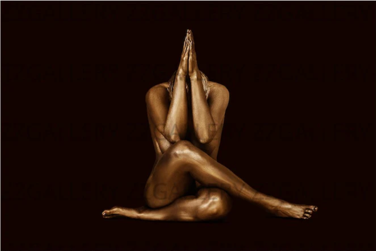 Feminine silhouette seated in gold body paint, cross-legged hands together on dark background by Zack Zublena.