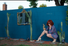 Surreal pop art photograph of a woman in a floral dress crouching in a garden with a golden cactus, framed by real cacti and a vintage mirror on a turquoise wall.