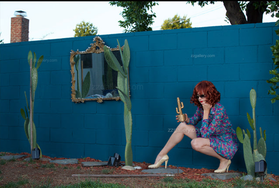 Surreal pop art photograph of a woman in a floral dress with a golden gun crouching by tall cacti against a turquoise wall, whimsical fine art wall décor by Zack Zublena.
