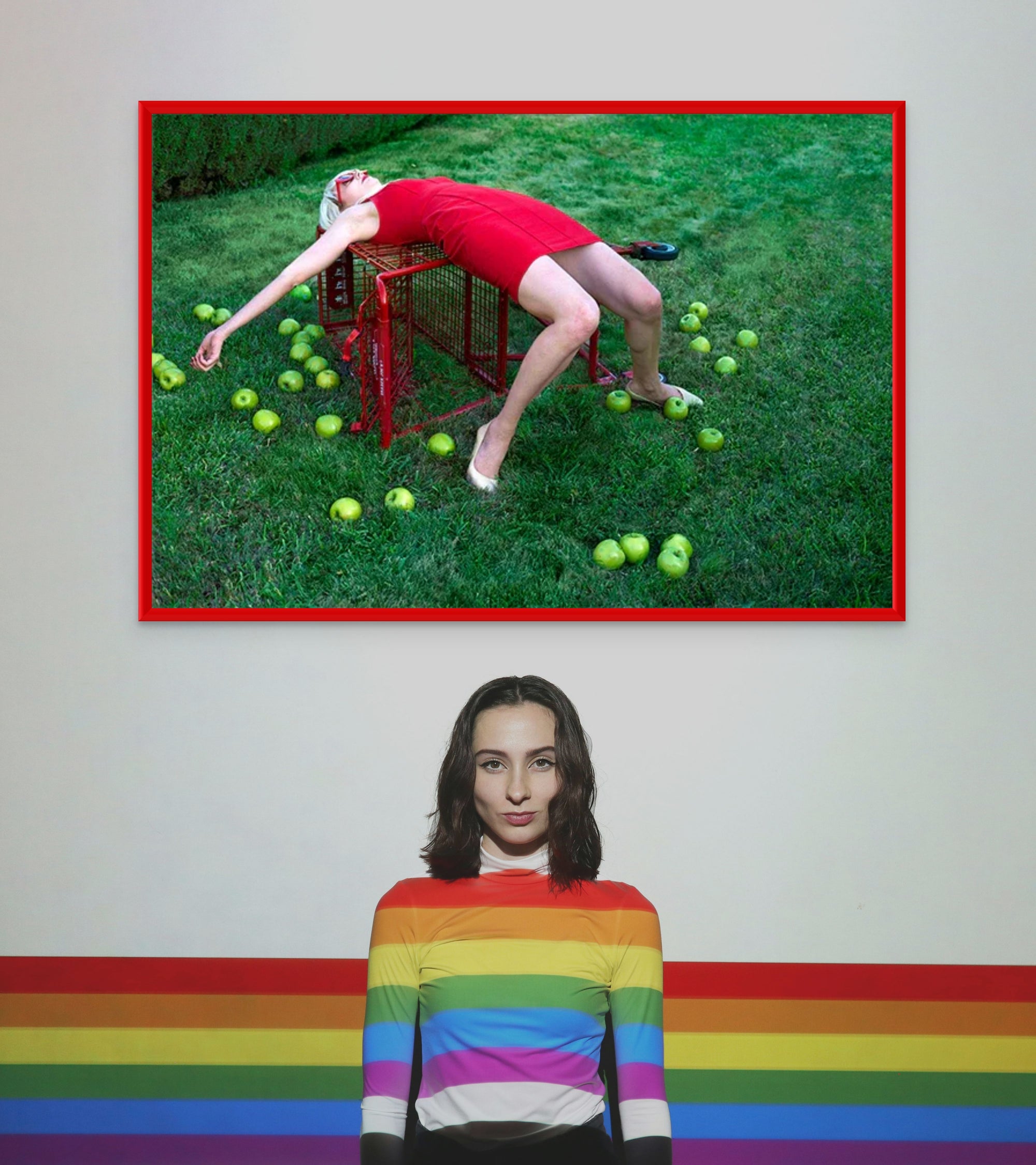 Surreal pop art photograph of a woman in a red dress stargazing while sprawled over a red shopping cart on grass, surrounded by apples – whimsical fine art print.
